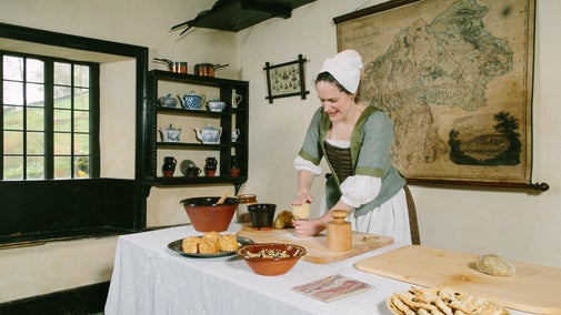 A woman in period costume creating food on a table in an old farmhouse kitchen